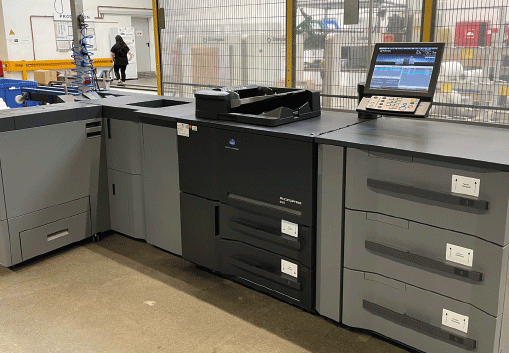 A large industrial printer with multiple trays and compartments is on a factory floor. A computer monitor with controls is attached, and there are shelves and workers in the background.
