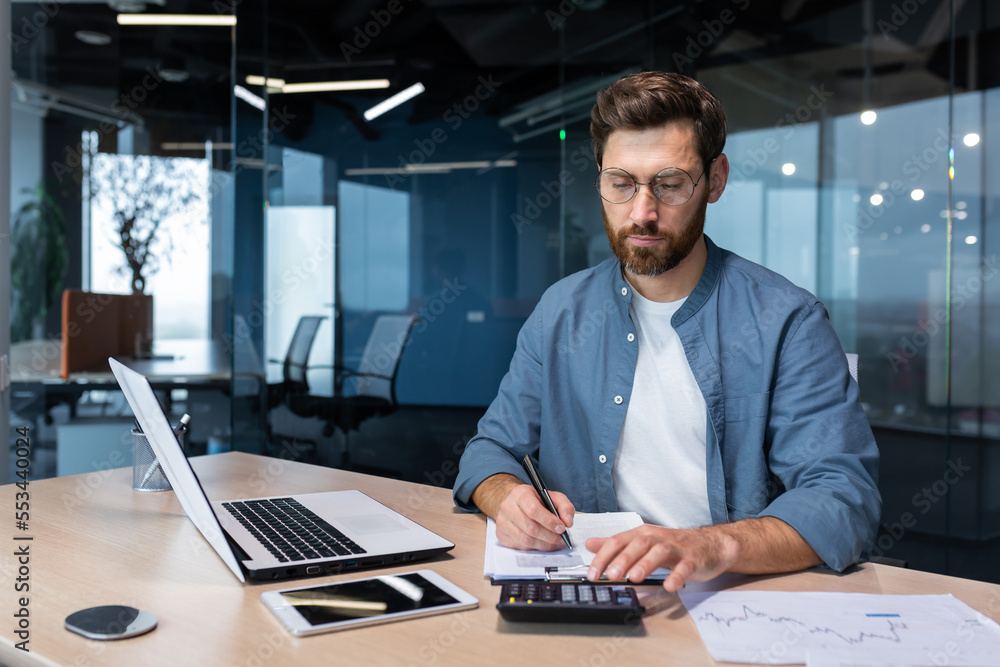 Ein Mann mit Brille arbeitet an einem Schreibtisch mit Laptop, Notebook, Taschenrechner und Smartphone in einem modernen Büro. Er schreibt Notizen, während er den Taschenrechner benutzt. Der Hintergrund zeigt Glaswände und einen leeren Konferenzraum.