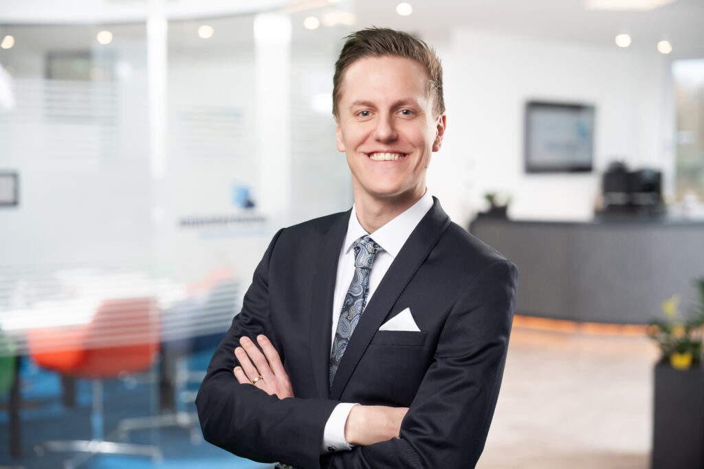 A young man in a dark suit and patterned tie stands confidently with arms crossed, smiling at the camera in a bright, modern office setting with frosted glass walls and contemporary furniture.