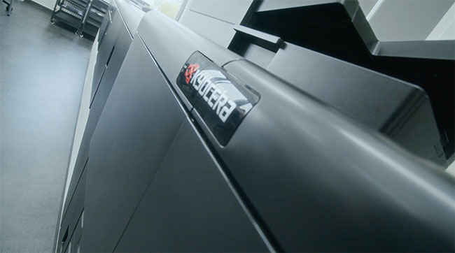 Nahaufnahme eines Kyocera-Bürodruckers oder -kopierers, die das Markenlogo auf der glatten, dunklen Oberfläche des Geräts in einem modernen, gut beleuchteten Arbeitsbereich zeigt.