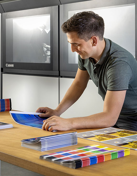 Ein Mann in einem grauen Polohemd begutachtet bunt bedruckte Blätter und Broschüren auf einem Holztisch vor großen Canon-Druckern.