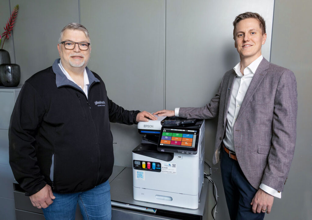 Zwei Männer stehen in einem Büro neben einem Epson-Drucker. Ein Mann trägt eine schwarze Jacke und eine Brille, der andere einen grauen Anzug. Beide lächeln und haben eine Hand auf dem Drucker.