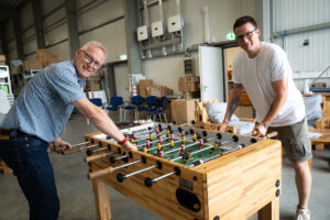 Zwei Männer, die lächelnd Tischfußball spielen, in einem geräumigen Raum im Industrial-Stil, der mit Kisten, Stühlen und Regalen gefüllt ist. Der Raum verfügt über große Fenster und einen Betonboden, was ihm eine ungezwungene Lagerhaus-Atmosphäre verleiht.