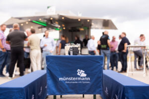 Eine blaue Tischdecke, auf der das Logo von „Münstermann – Dokumente mit System“ im Vordergrund zu sehen ist, während sich im unscharfen Hintergrund eine Gruppe von Menschen in der Nähe eines Imbissstandes im Freien unterhält.