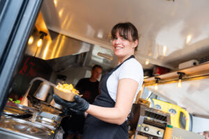 Eine lächelnde Frau, die eine Schürze und schwarze Handschuhe trägt, serviert Essen aus einem Imbisswagen und hält dabei eine Schüssel mit fertig zubereiteten Speisen in der Hand. Der Innenraum des Imbisswagens ist zu sehen; im Hintergrund sind eine weitere Person und verschiedene Küchengeräte zu erkennen.