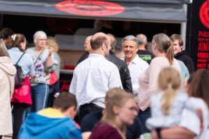 Eine Gruppe von Menschen steht vor einem Imbissstand bei einer Veranstaltung im Freien und unterhält sich. Einige lächeln und unterhalten sich, und die Szene wirkt lebhaft, da im Hintergrund viele Menschen zu sehen sind.