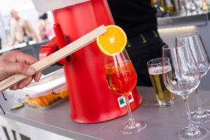 Im Hintergrund ist eine Hand zu sehen, die einen Holzstab hält, an dem ein roter Getränkespender befestigt ist. Auf der Theke steht ein Weinglas, gefüllt mit einem orangefarbenen Cocktail und garniert mit einer Orangenscheibe, neben leeren Gläsern und einem Bierglas.