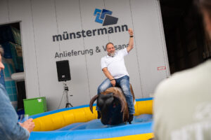 Ein Mann in einem weißen Hemd reitet auf einem mechanischen Bullen, lächelt und hält einen Arm in die Höhe, vor einem Gebäude mit der Aufschrift „Münstermann Anlieferung & Versand“. Im Vordergrund schauen Menschen zu.