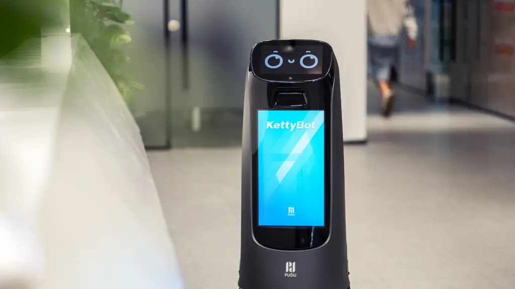 Ein Pudu KettyBot-Serviceroboter mit einem digitalen Gesicht auf seinem Bildschirm steht in einem Büroflur.