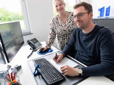 Ein Mann sitzt an einem Schreibtisch mit Tastatur, Monitor und Telefon und schreibt etwas auf ein Blatt Papier, während eine Frau lächelnd neben ihm steht. Beide befinden sich in einem hellen Büro mit Fenstern und blauen Auszeichnungen, die in einem Regal ausgestellt sind.